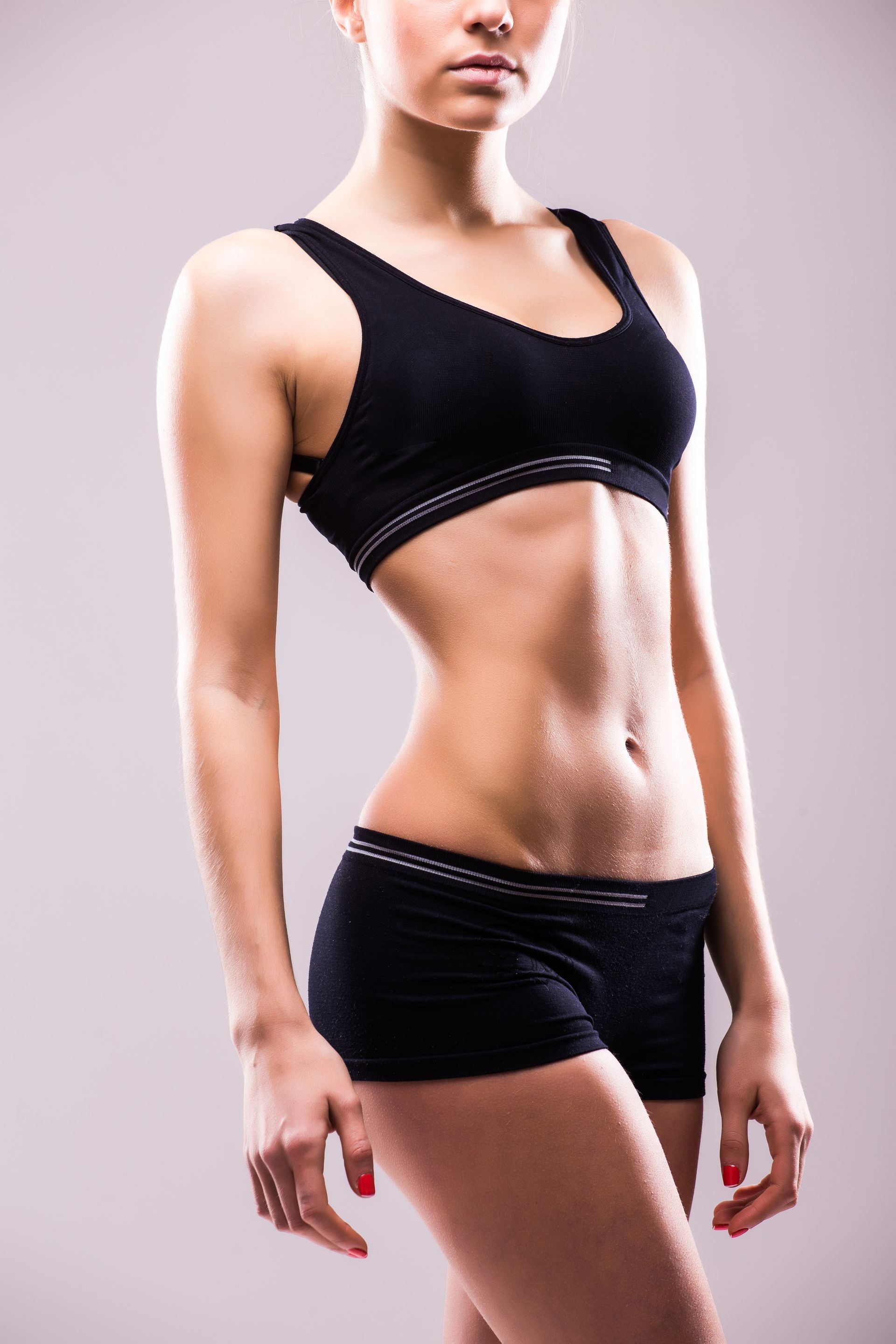 A woman in a sports bra and shorts is standing in front of a gray background.