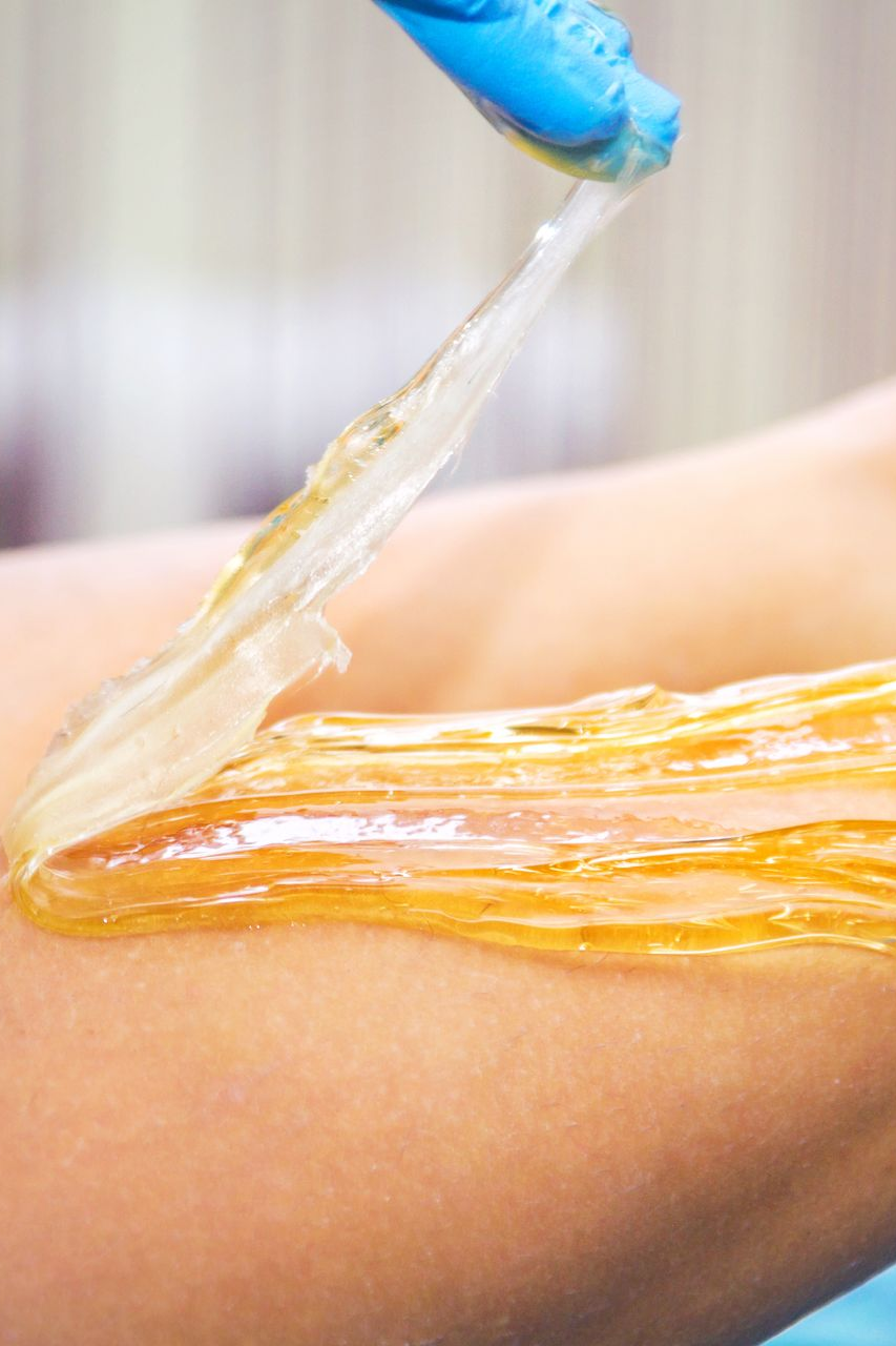 Hand in blue glove applying amber-colored sugaring paste to an arm.