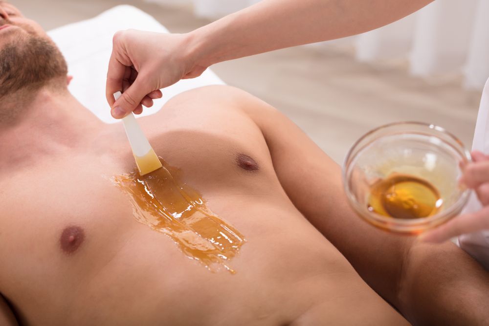 Man having chest waxed. Technician applies wax with a spatula; bowl of wax present.
