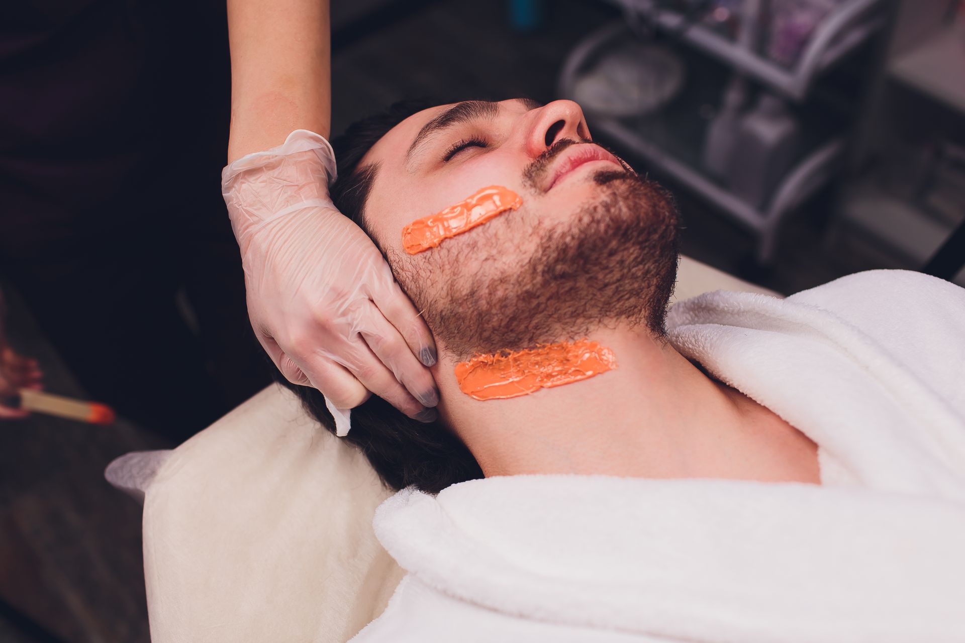 Man getting facial wax with orange wax strips on face and neck in spa.