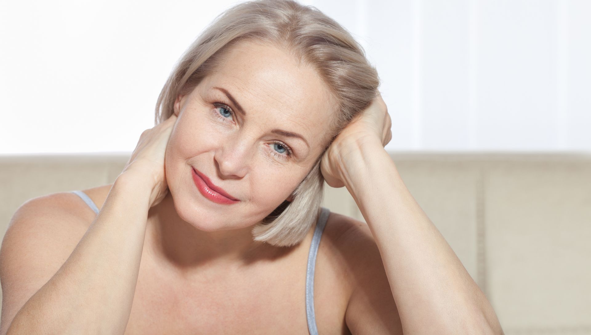 Woman with short blonde hair smiles, resting head on hands. Light-colored tank top.