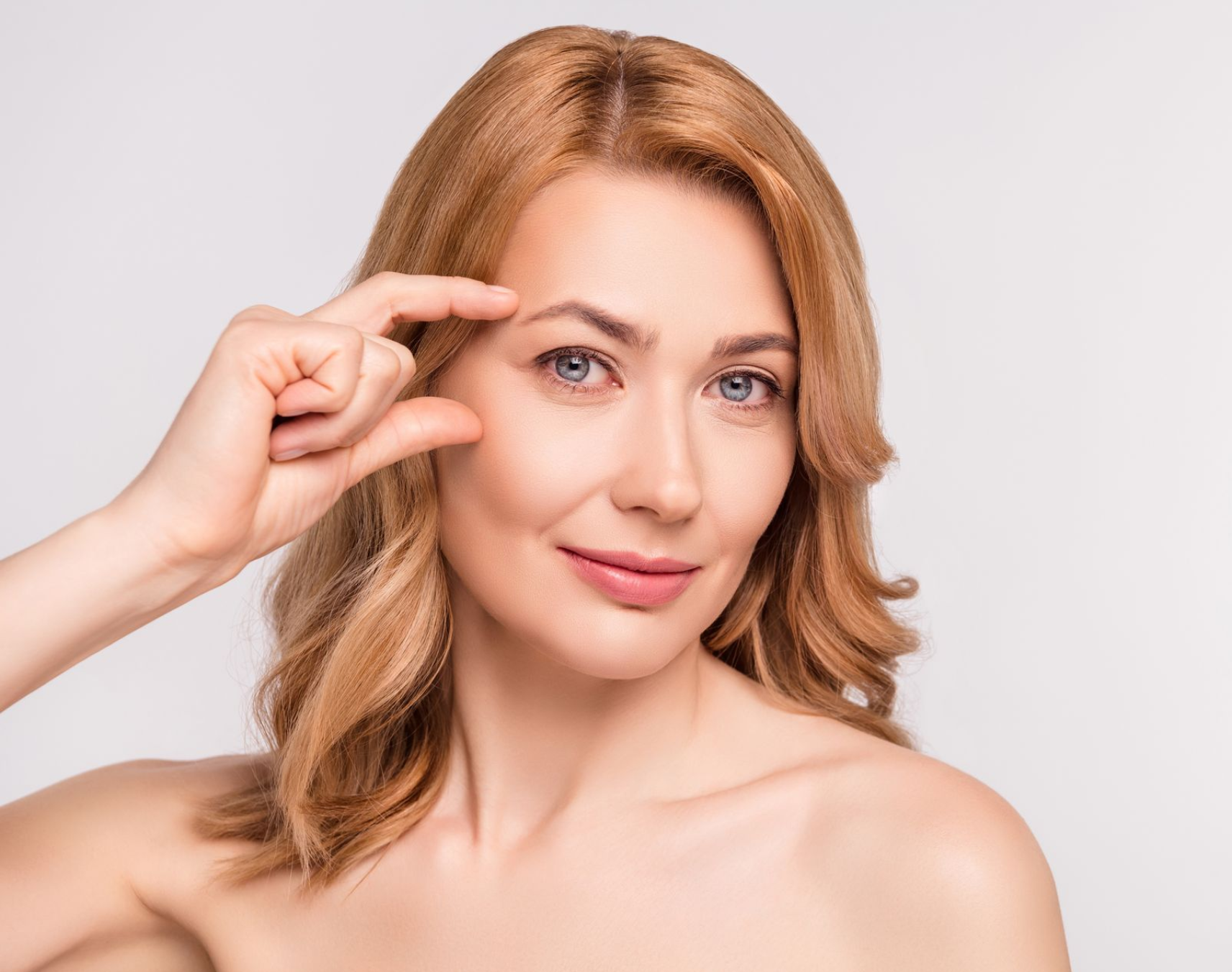 Woman holding fingers near her eyebrow, smiling, with wavy red-blonde hair.
