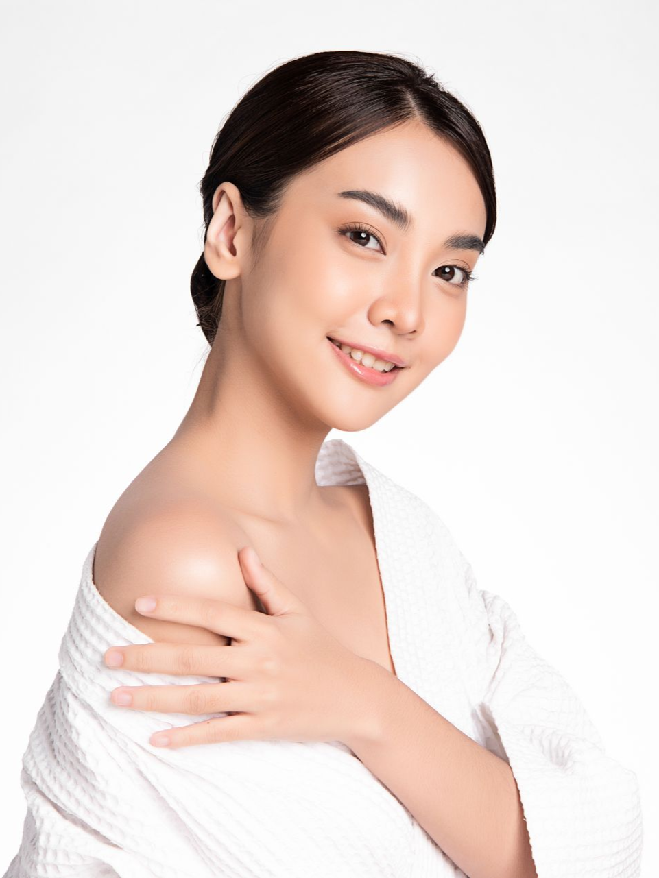 Woman in white robe, smiles, touches her shoulder, soft lighting.