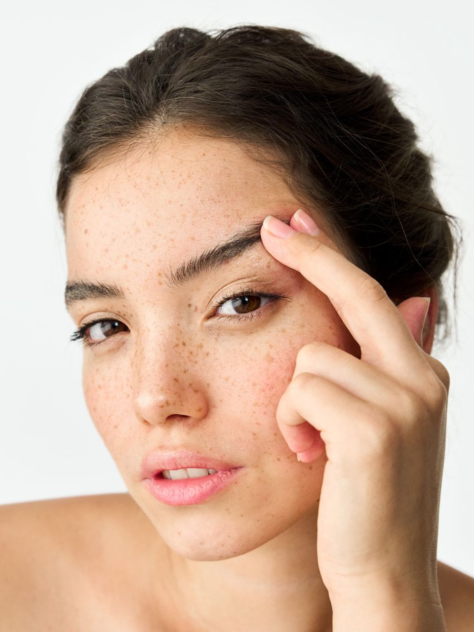 Woman touching eyebrow, freckled face, brown eyes, light skin, neutral background.