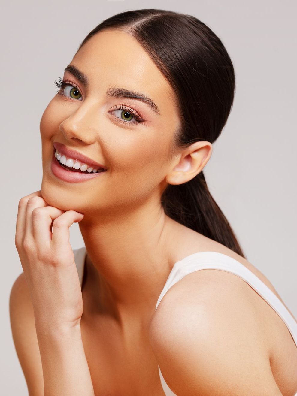 A woman in a white tank top is smiling and touching her face.