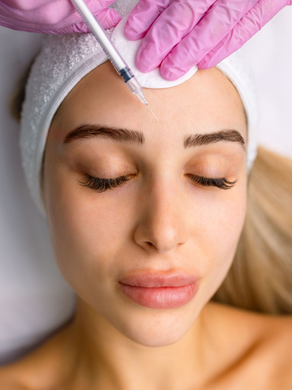 Person receiving facial injection, pink gloved hand with syringe, cotton swab on forehead.