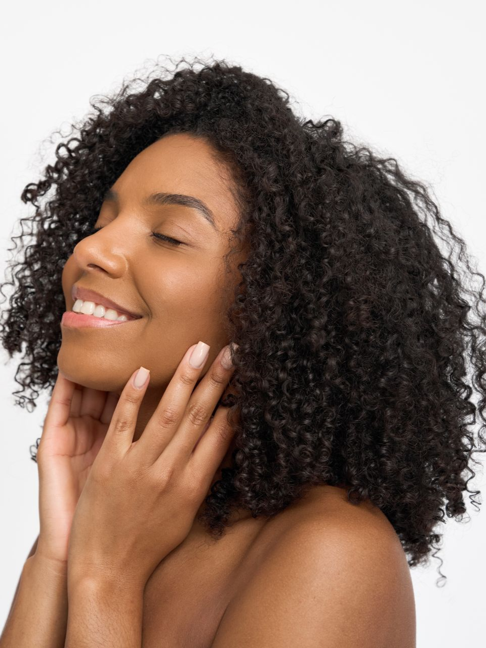 Woman with curly hair smiles, eyes closed, hands gently touching her face.