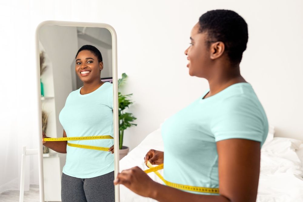 Woman measuring waist with a tape in front of a mirror, smiling. Pale blue shirt, gray pants.