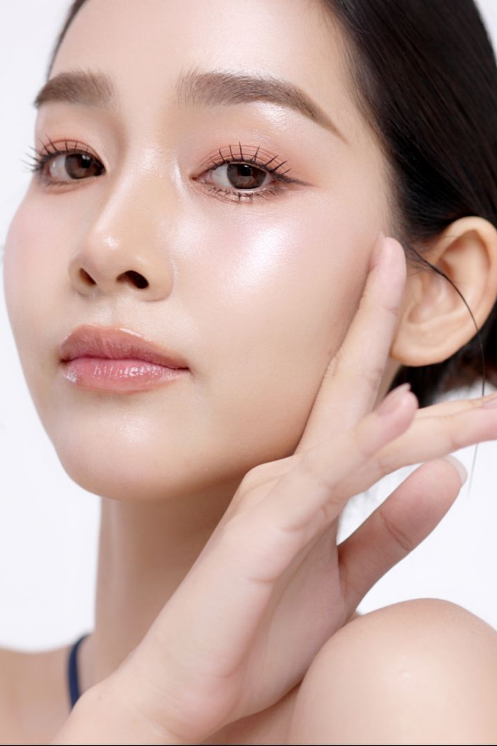 Woman with glowing skin, touching her face. She wears light pink eye shadow, against a white background.