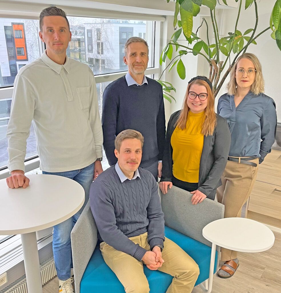 Five colleagues pose in a modern office setting. The group includes two men and three women.