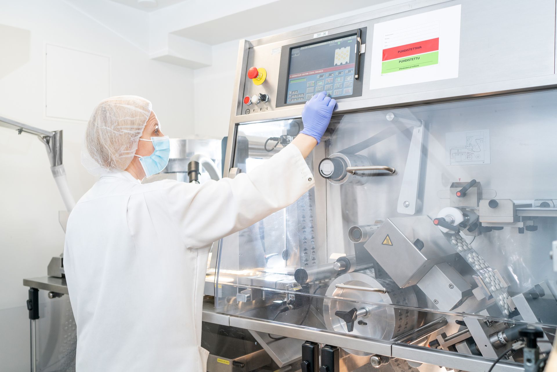Woman in lab coat and mask operates machinery in a pharmaceutical manufacturing setting.