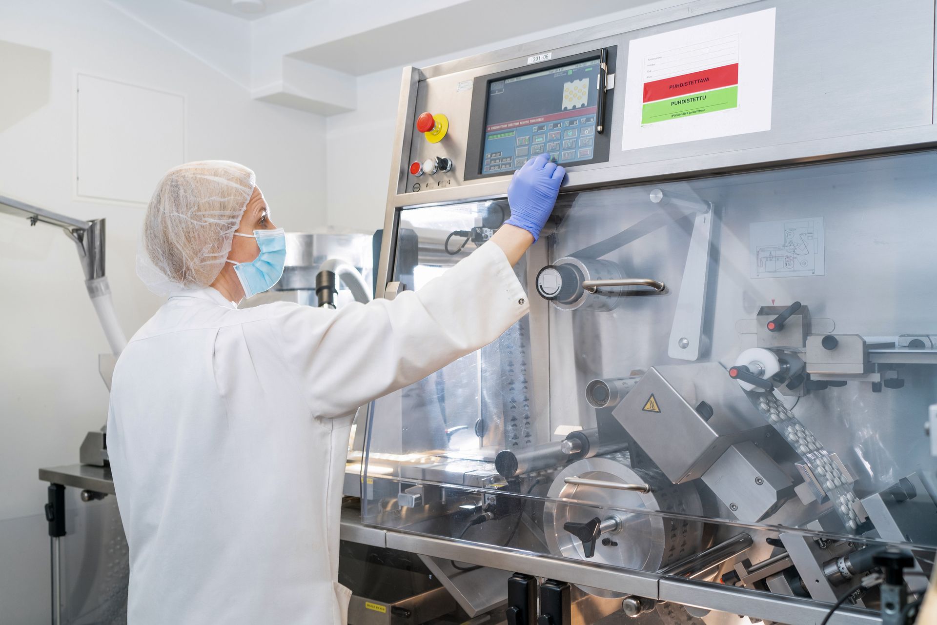 Woman in lab coat and mask operates machinery in a pharmaceutical manufacturing setting.