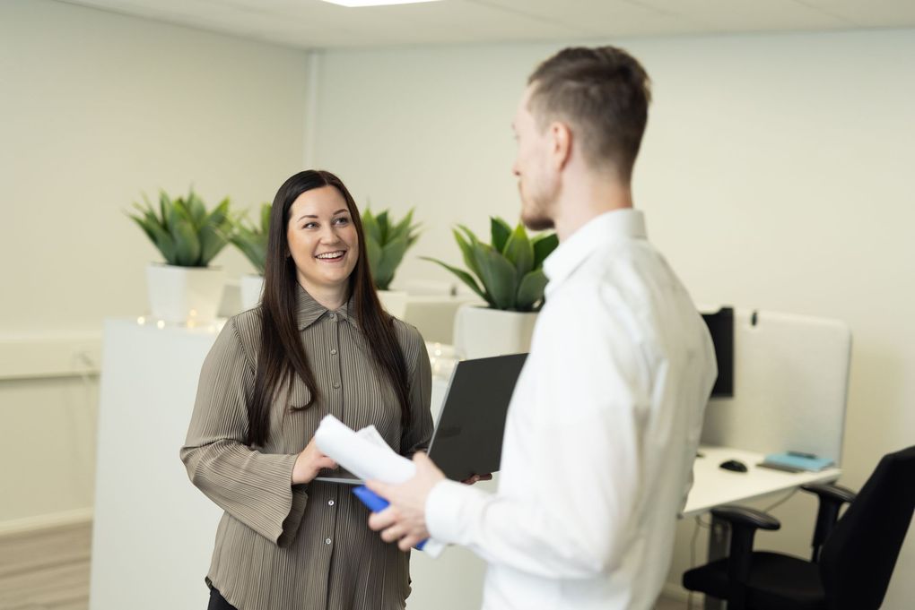 Woman smiling, talking to a man holding papers in a modern office with potted plants.