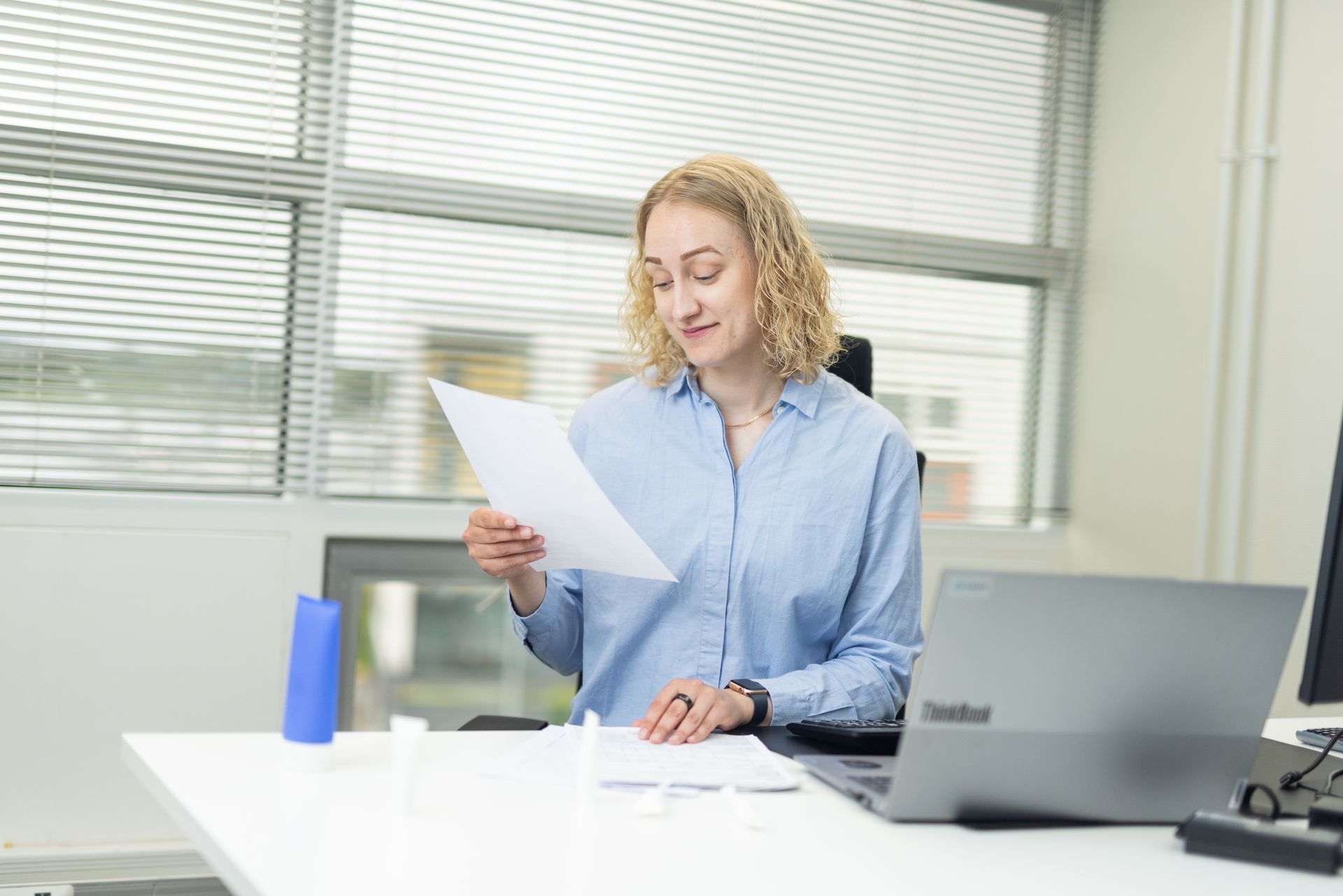 Woman with blonde curly hair, reading a paper at a desk in an office, looking down with a slight smile.