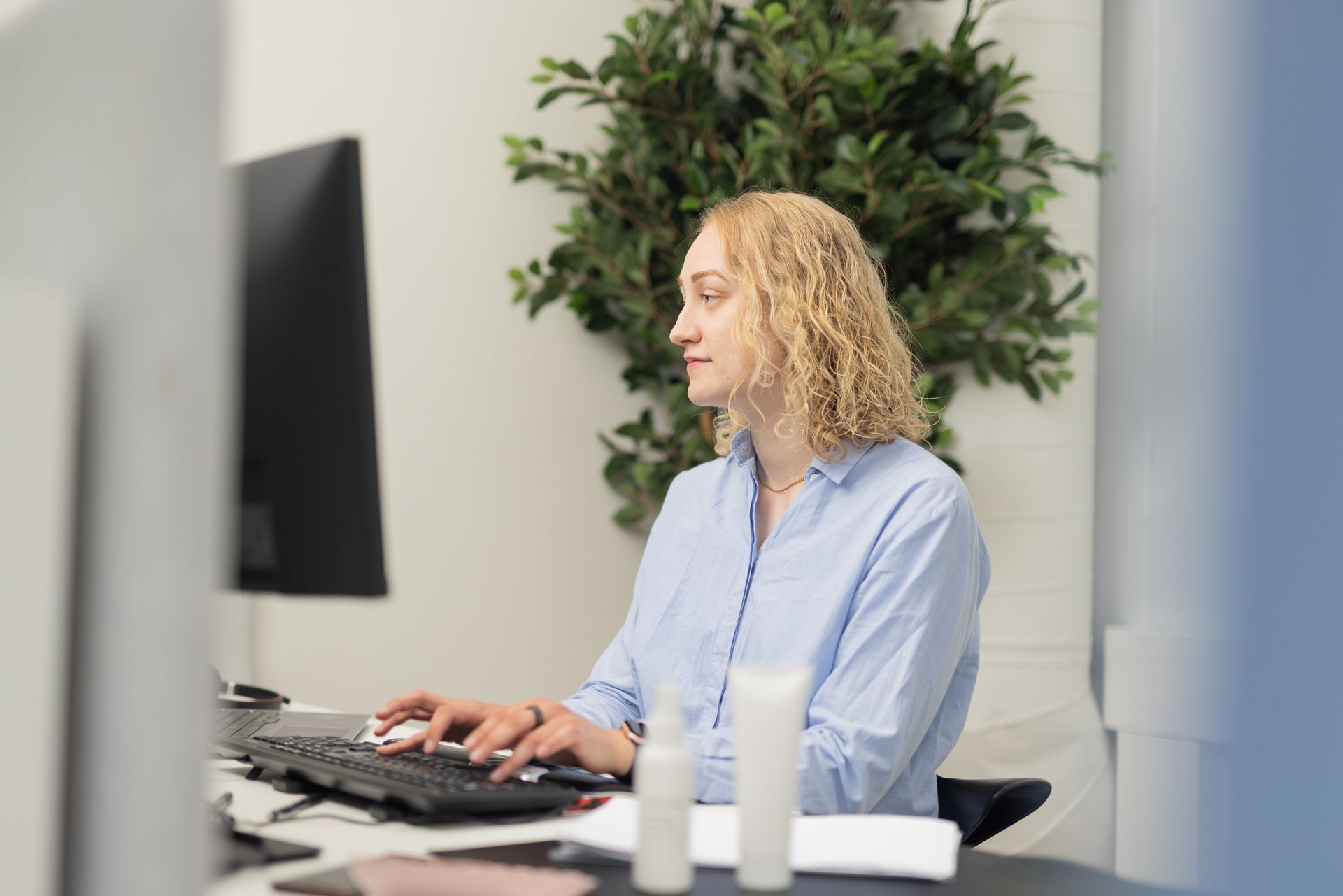 Woman with curly blonde hair typing at a computer in an office setting, with a green plant in the background.