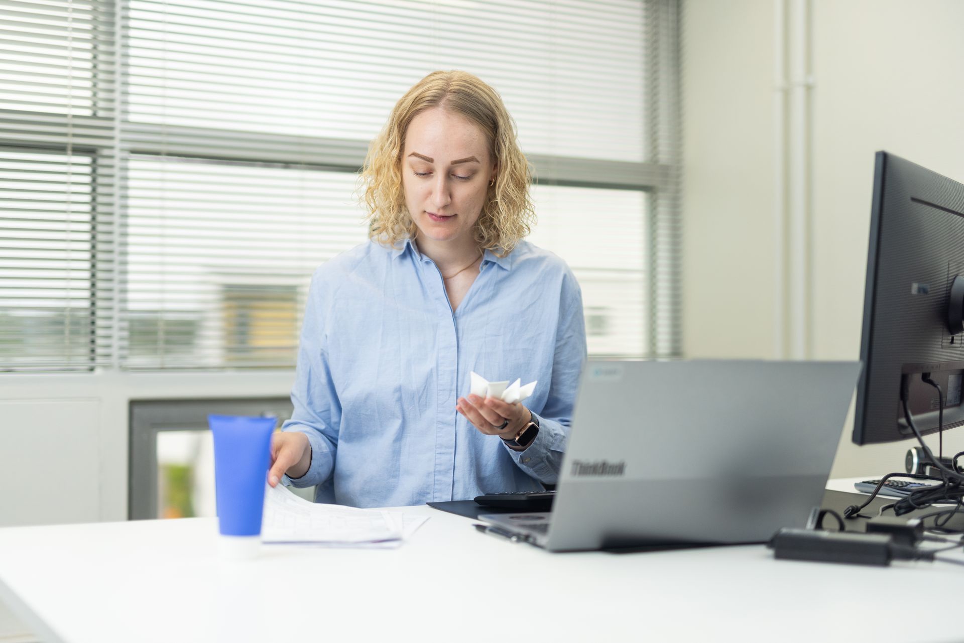 Woman at a desk with a laptop and papers; holds crumpled paper, blue cup. Bright office setting.