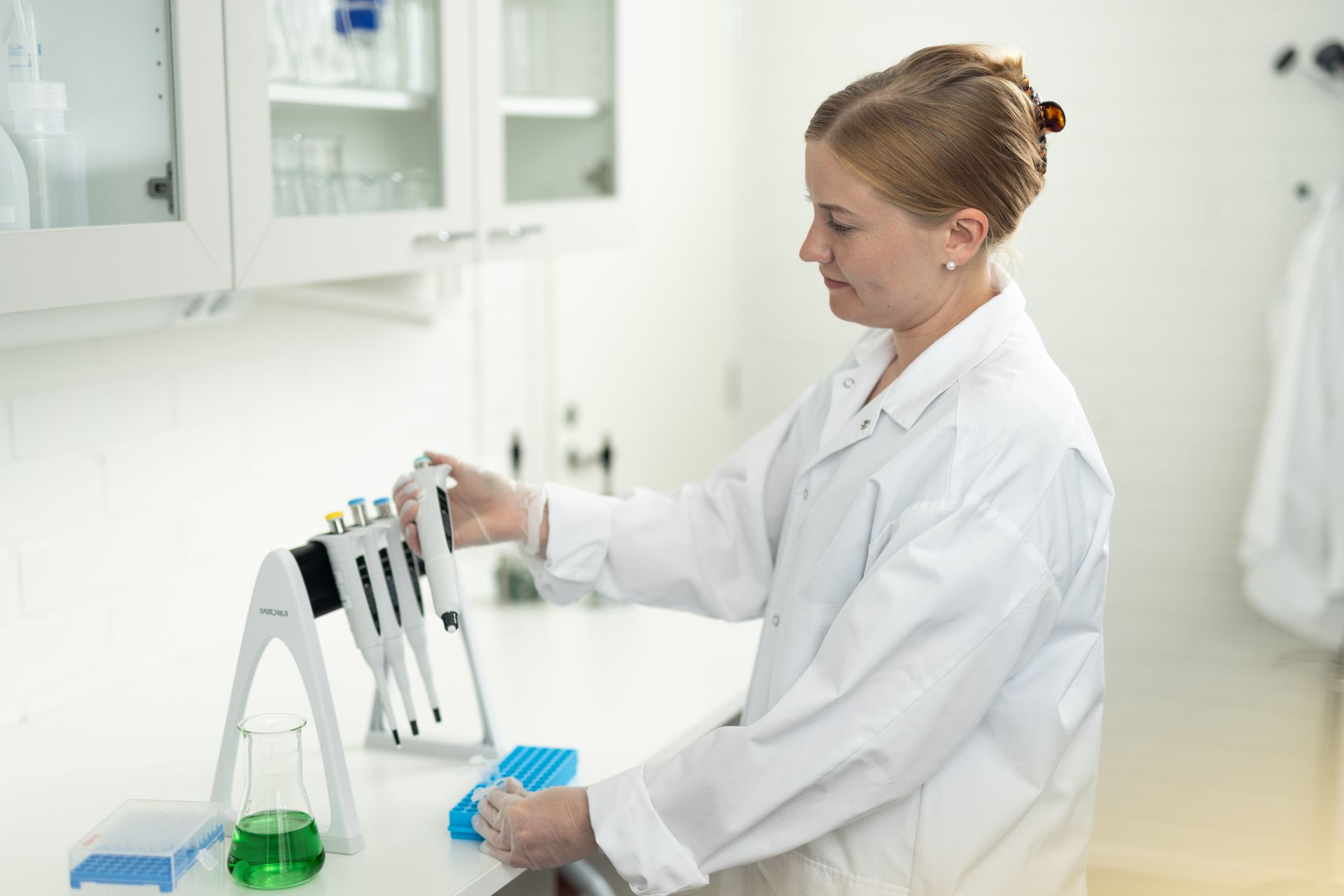Woman in lab coat uses a pipette in a lab, with glassware and equipment in a clean, white environment.