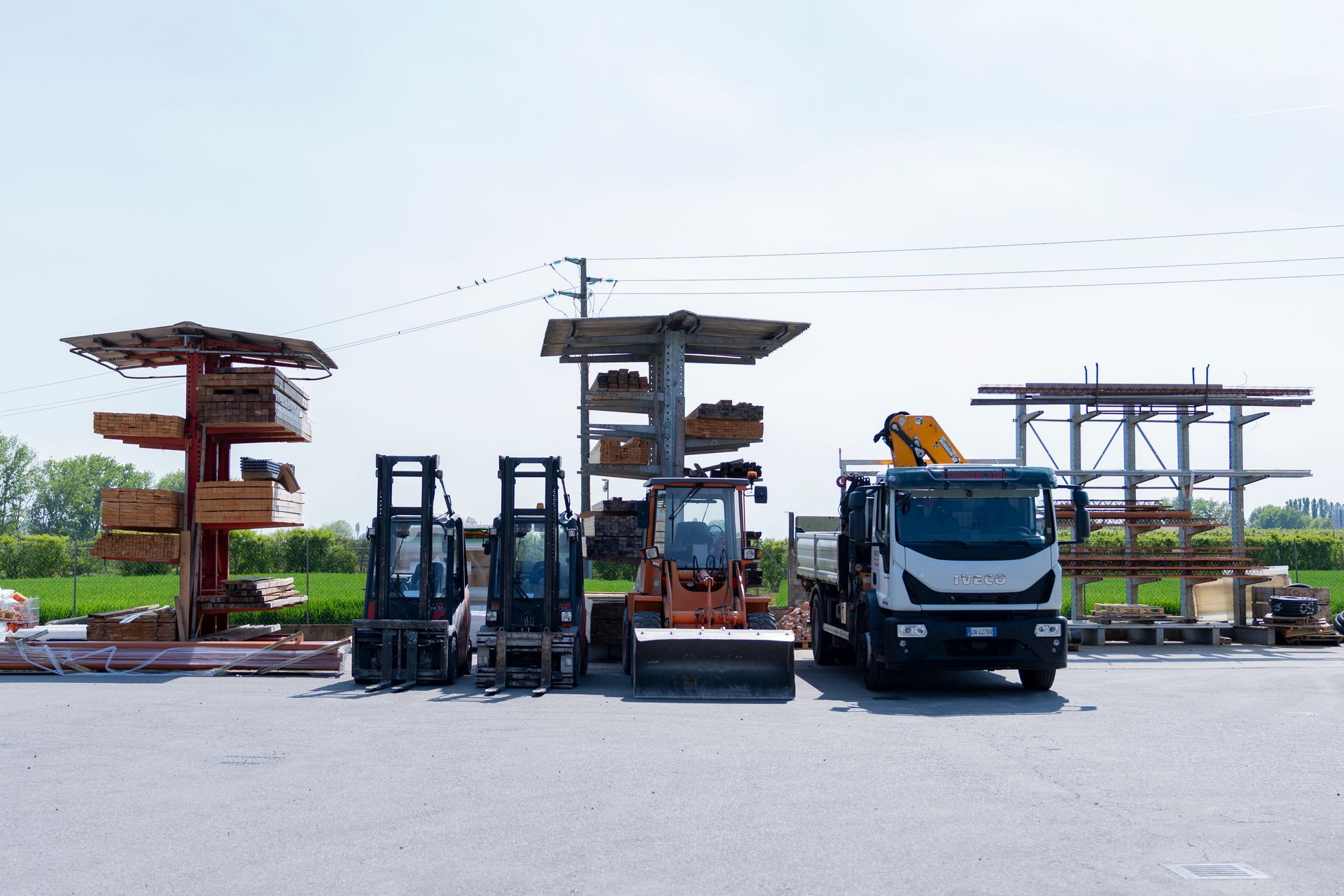In un piazzale sono parcheggiati carrelli elevatori e un camion, con scaffalature in legno per pallet e linee elettriche sullo sfondo.