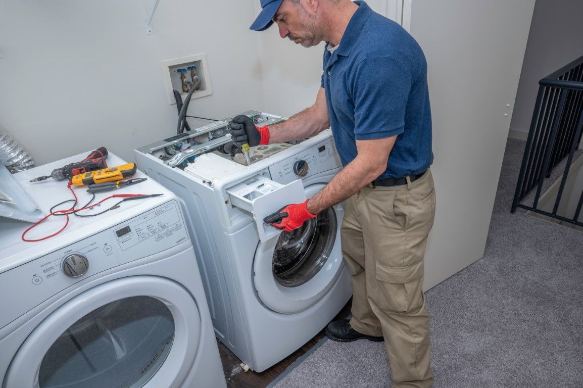 Man in blue shirt and gloves repairing a washing machine in a utility room.