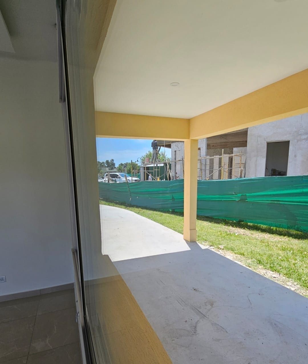 Patio cubierto con columnas amarillas, suelo de hormigón y valla de construcción verde en el exterior.