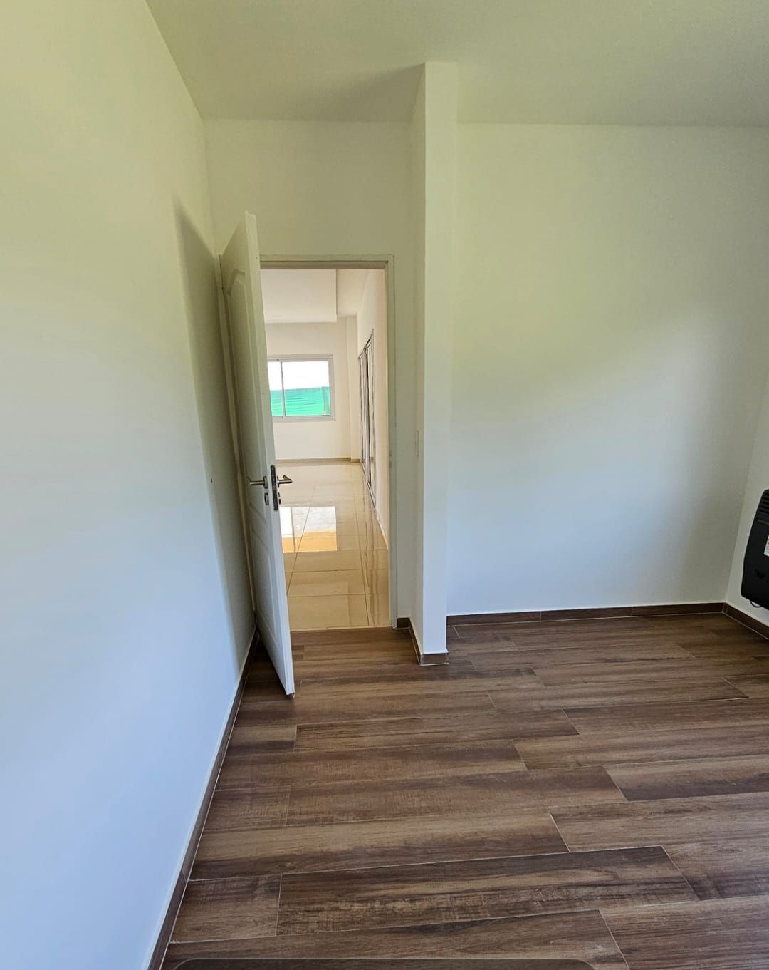 Habitación vacía con suelo de madera, puerta abierta y un pasillo luminoso al fondo.