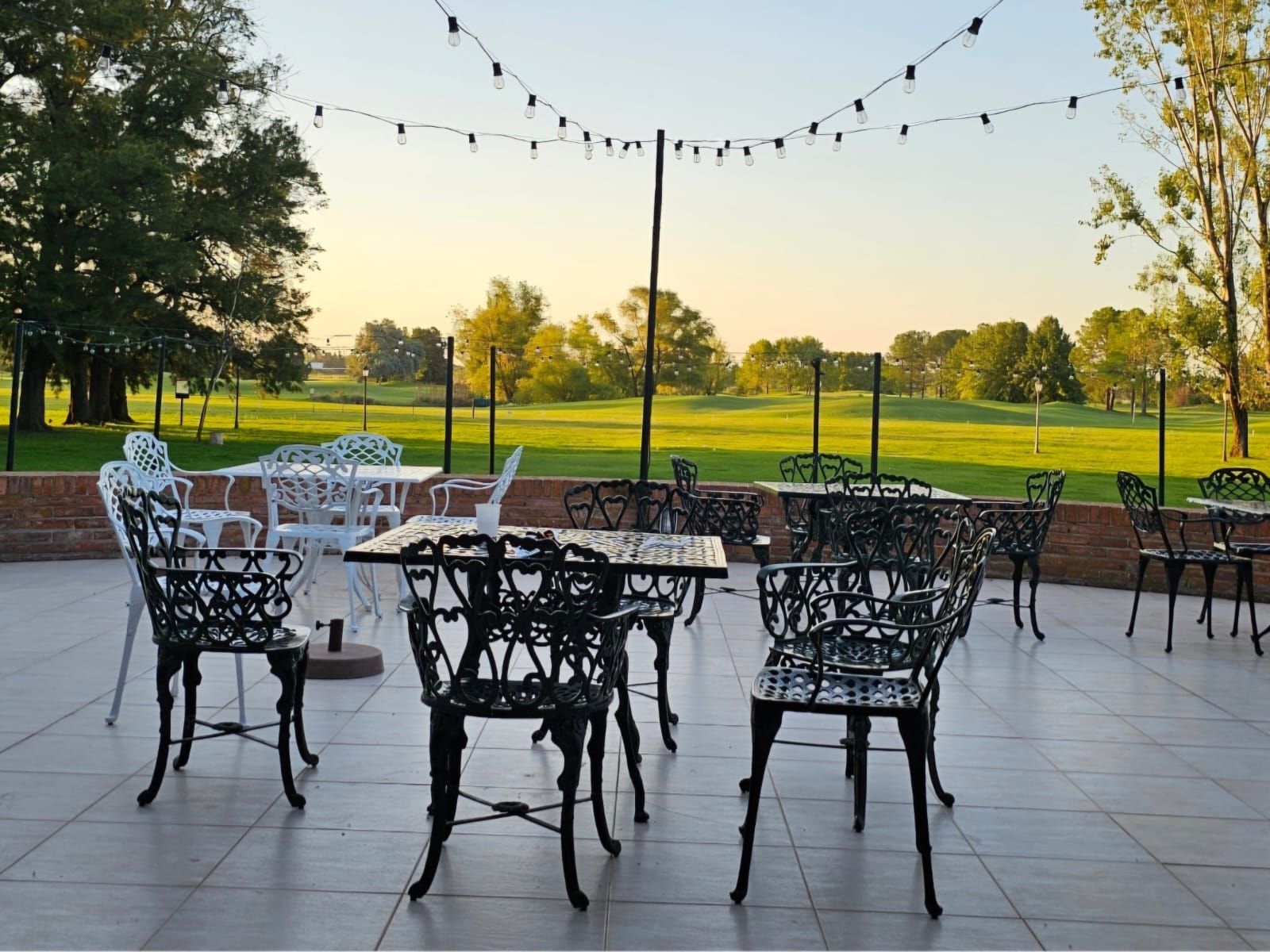 Patio exterior con sillas y mesas de metal negro bajo guirnaldas de luces junto a un parque verde al atardecer.