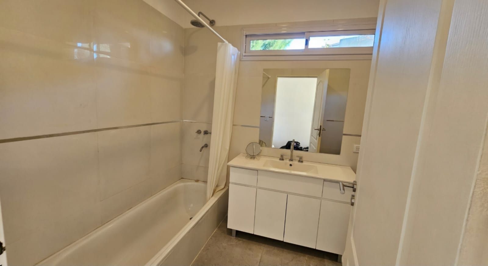 Baño luminoso con paredes alicatadas en color beige, bañera a la izquierda y tocador blanco con espejo al fondo.