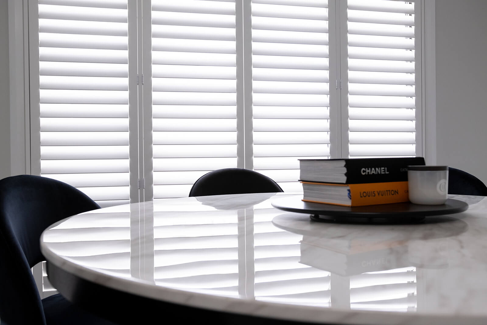 A Stack of Books Sits on a Table in Front of a Window With White Shutters — Abode Shutters & Blinds In Taree South, NSW