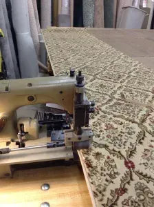 A sewing machine stitching the edge of a patterned rug in a workshop setting.