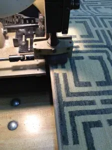 A rug being sewn by a large machine in a workshop; the rug has a geometric pattern.