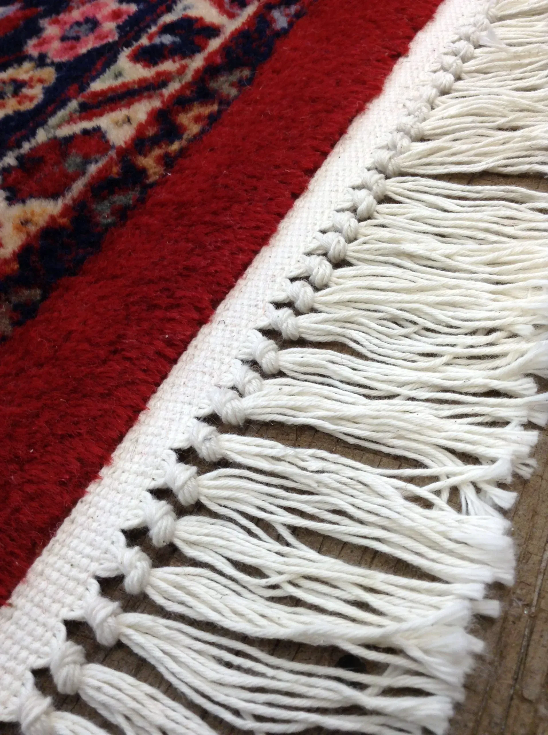 Close-up of a red rug edge with white fringe knotted at the edge. Part of a patterned rug visible.