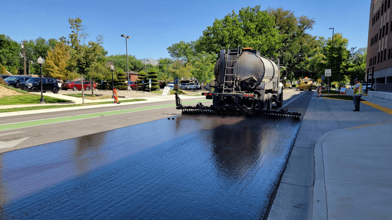 Truck spraying a glossy black sealcoat across a wide road surface to protect and extend pavement life during an asphalt paving service in Arvada, CO