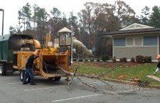 Tractor Mounted for Cutting a Tree — Williamsburg, VA — Top Notch Tree Service Man feeding branches into a wood chipper near a playground and building.