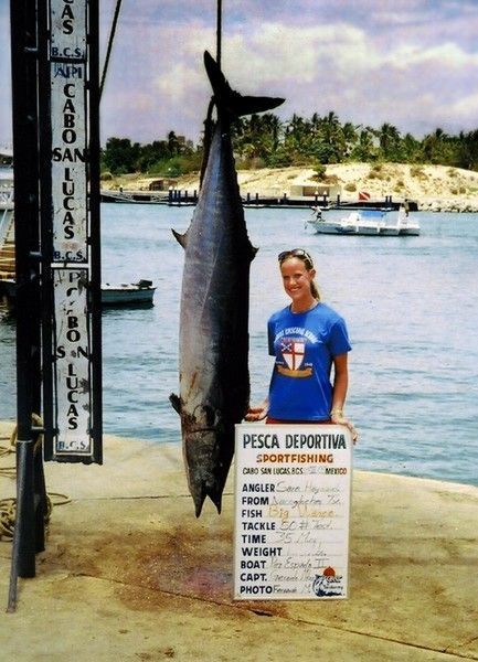 A woman in a blue shirt holds a large fish in front of a sign that says pesca deportiva