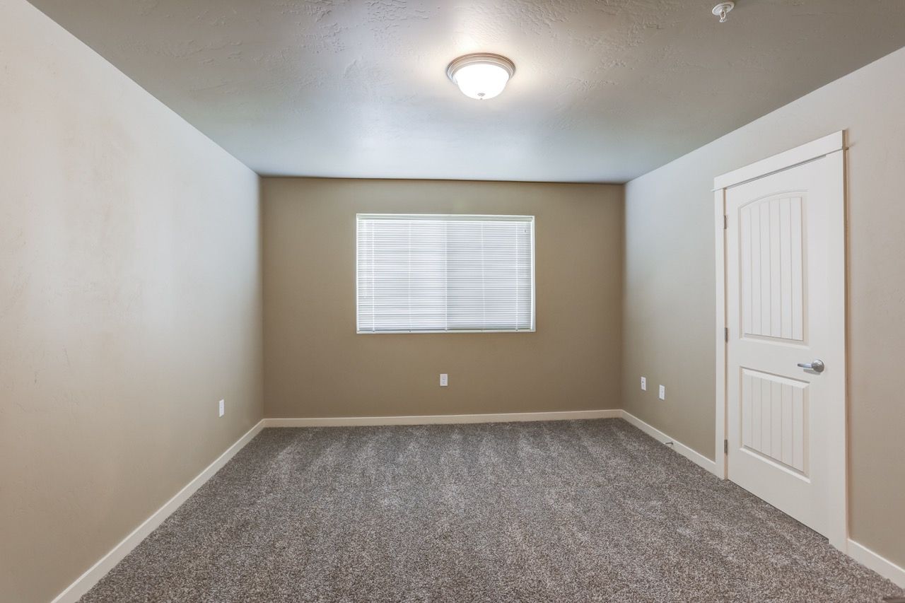 Small empty beige bedroom with a window and a white door, carpeted floor.