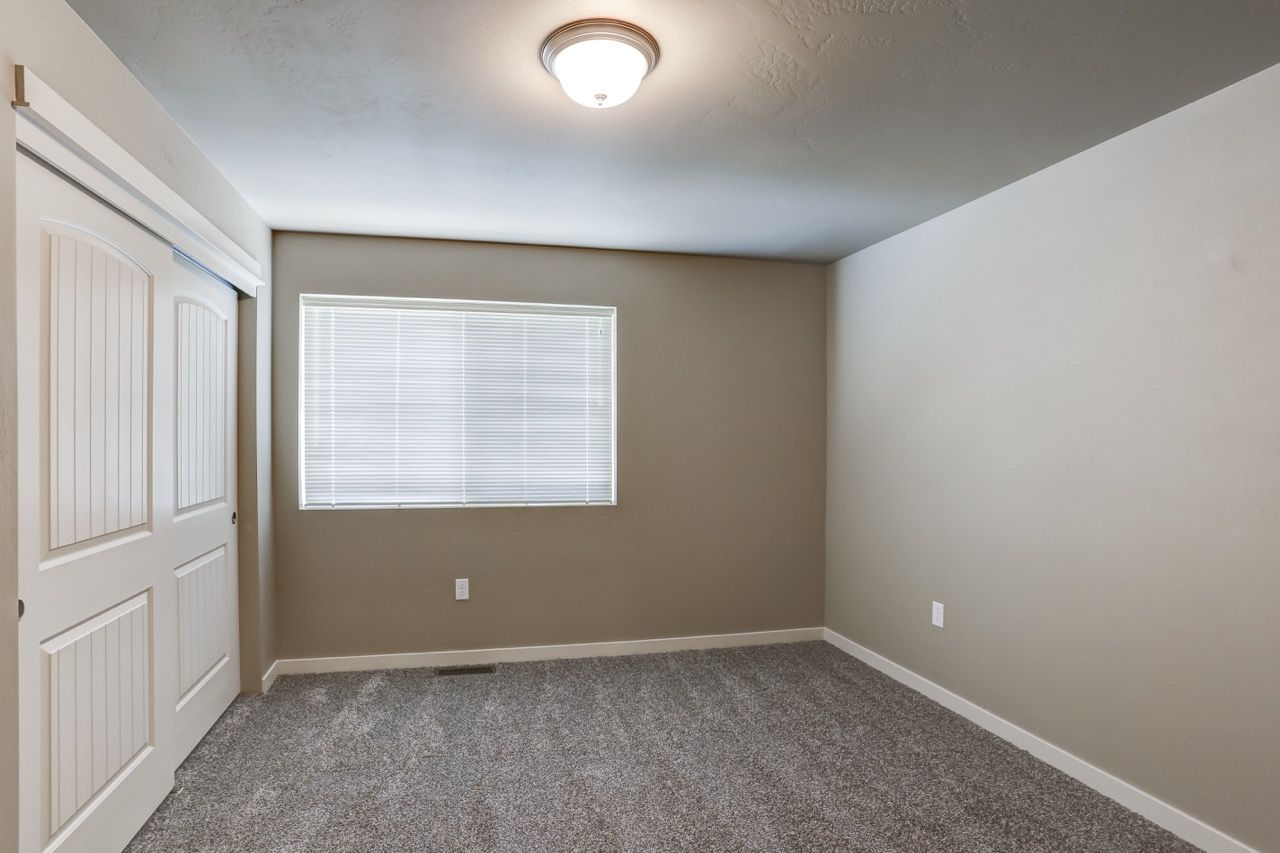 Empty bedroom with beige walls, white closet doors, window with blinds, and carpet.