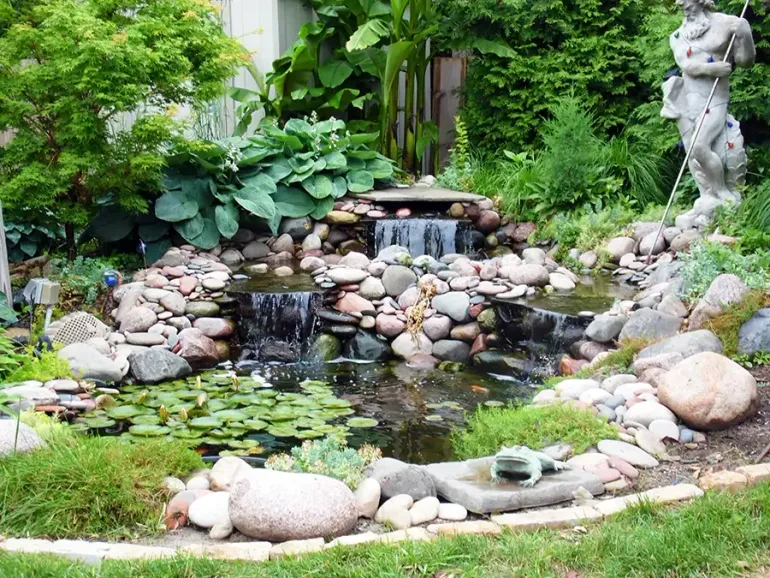 Stone waterfall pond in a lush garden with lily pads and a statue on the right