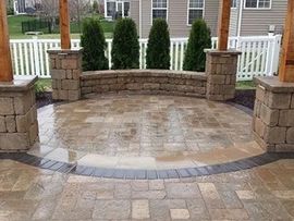 Stone patio with curved retaining wall, brick columns, and white fence in a backyard