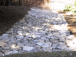 Stone-paved path in sunlight, bordered by rough ground and a wooden fence.