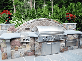 Built-in outdoor stone grill with stainless steel lids and red flower planters in a landscaped patio