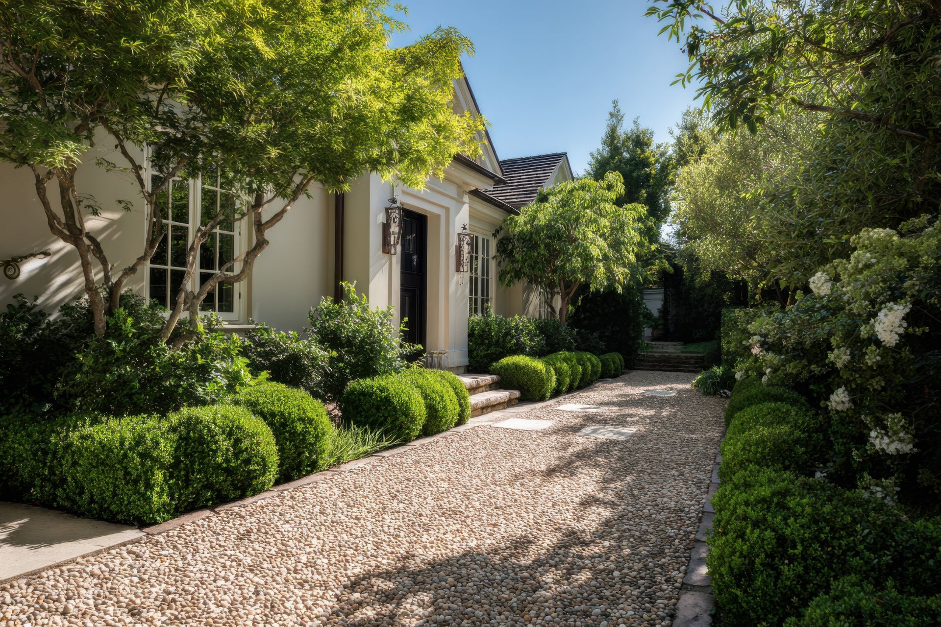 Sunlit garden path beside a house, lined with lush green shrubs and trees