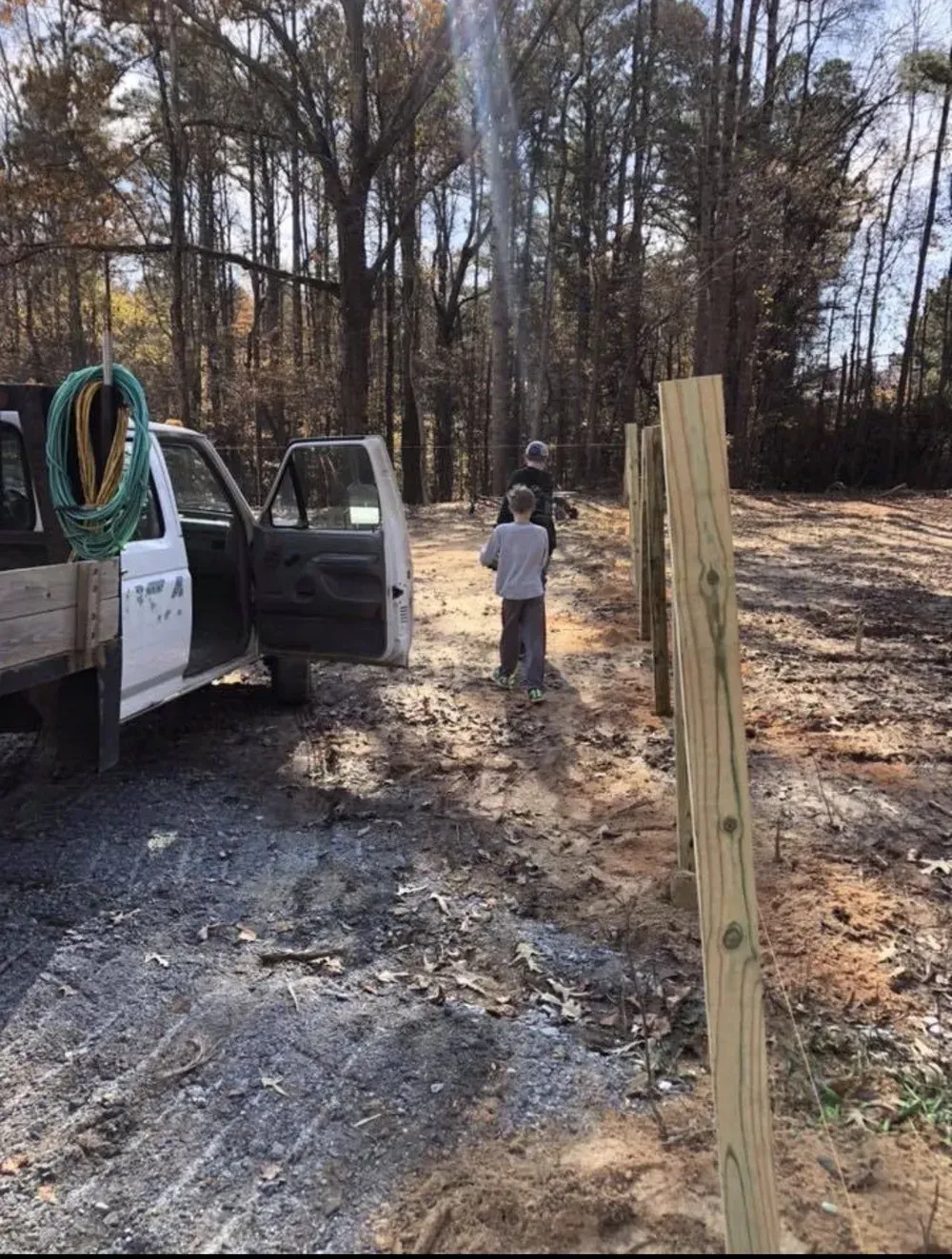 Worker by pickup truck at wooded construction site with upright wooden posts and gravel ground