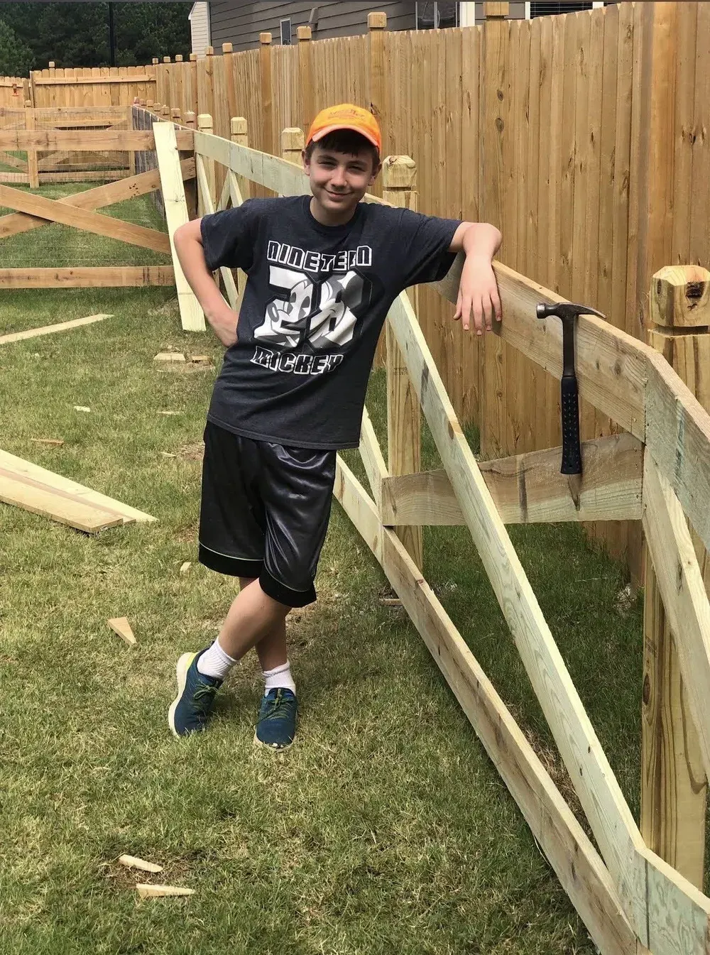 Person leaning on a wooden fence in a backyard, wearing a black T-shirt, shorts, and an orange cap.