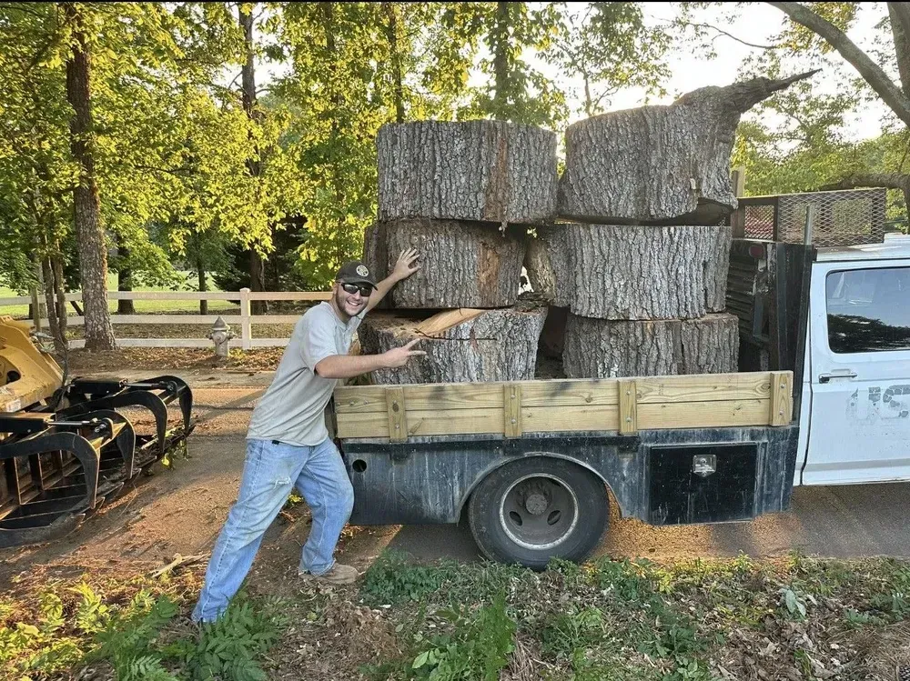 Man loading large stacked hay bales onto a pickup truck in a wooded yard
