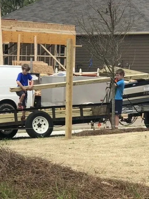 Two people loading boards onto a trailer in a driveway, with a white van and house under construction nearby