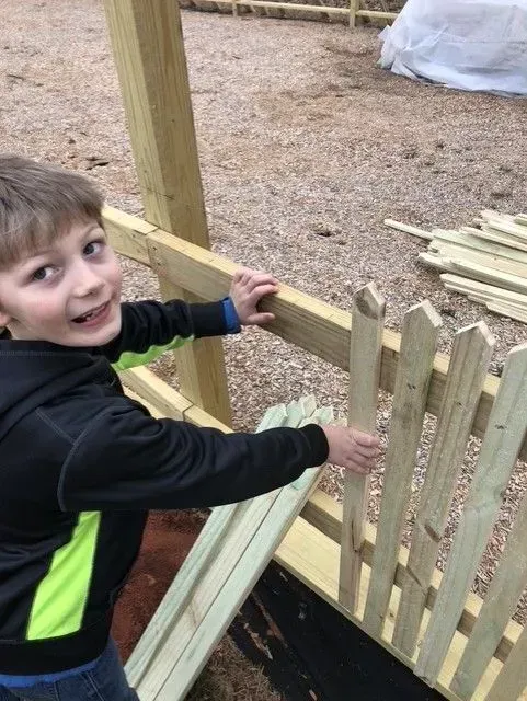 Smiling child in a black and neon green jacket opening a wooden gate outdoors