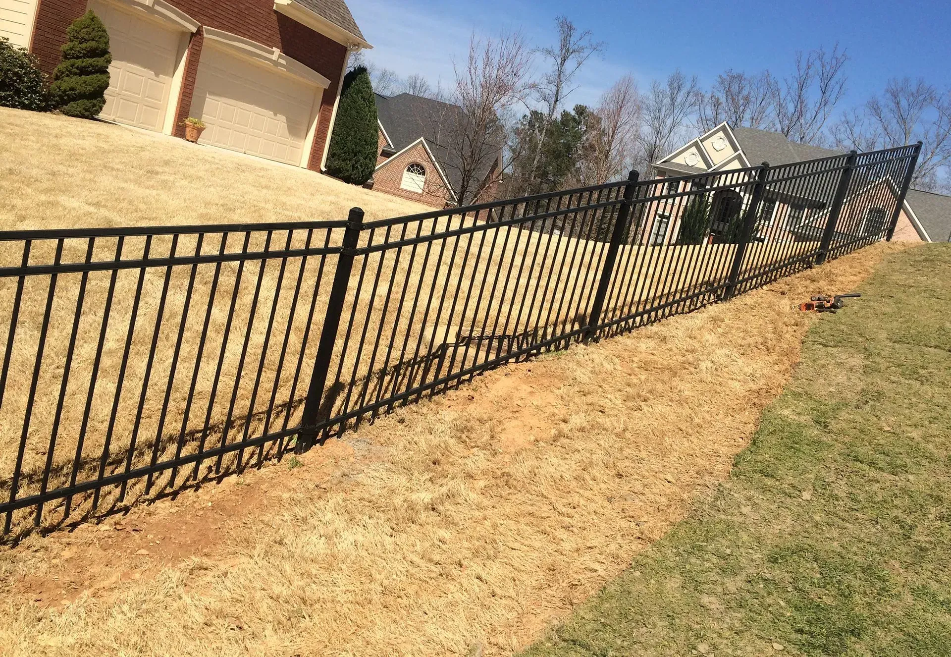 Black metal fence along a sloped tan lawn beside suburban houses on a sunny day