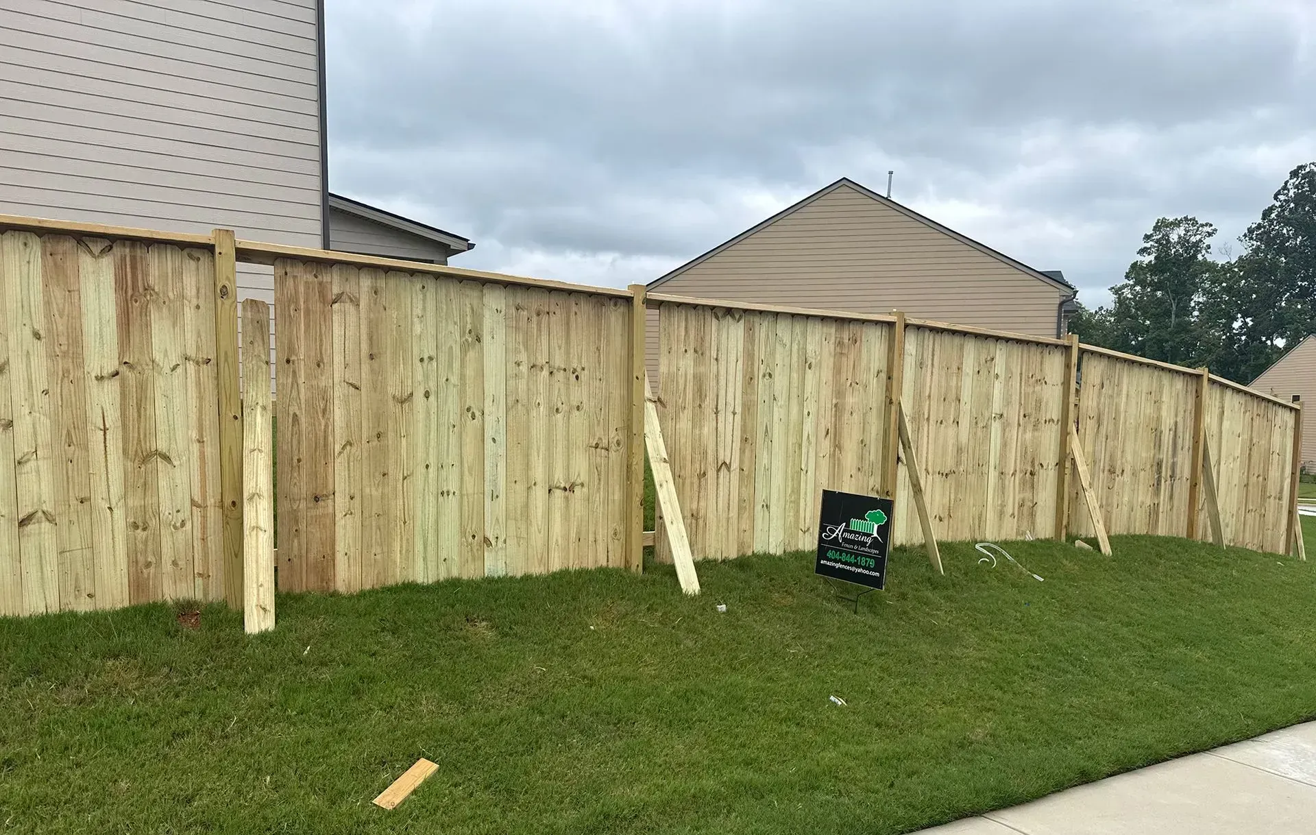 New wooden privacy fence along a grassy yard beside a driveway, with a fence post leaning against it.