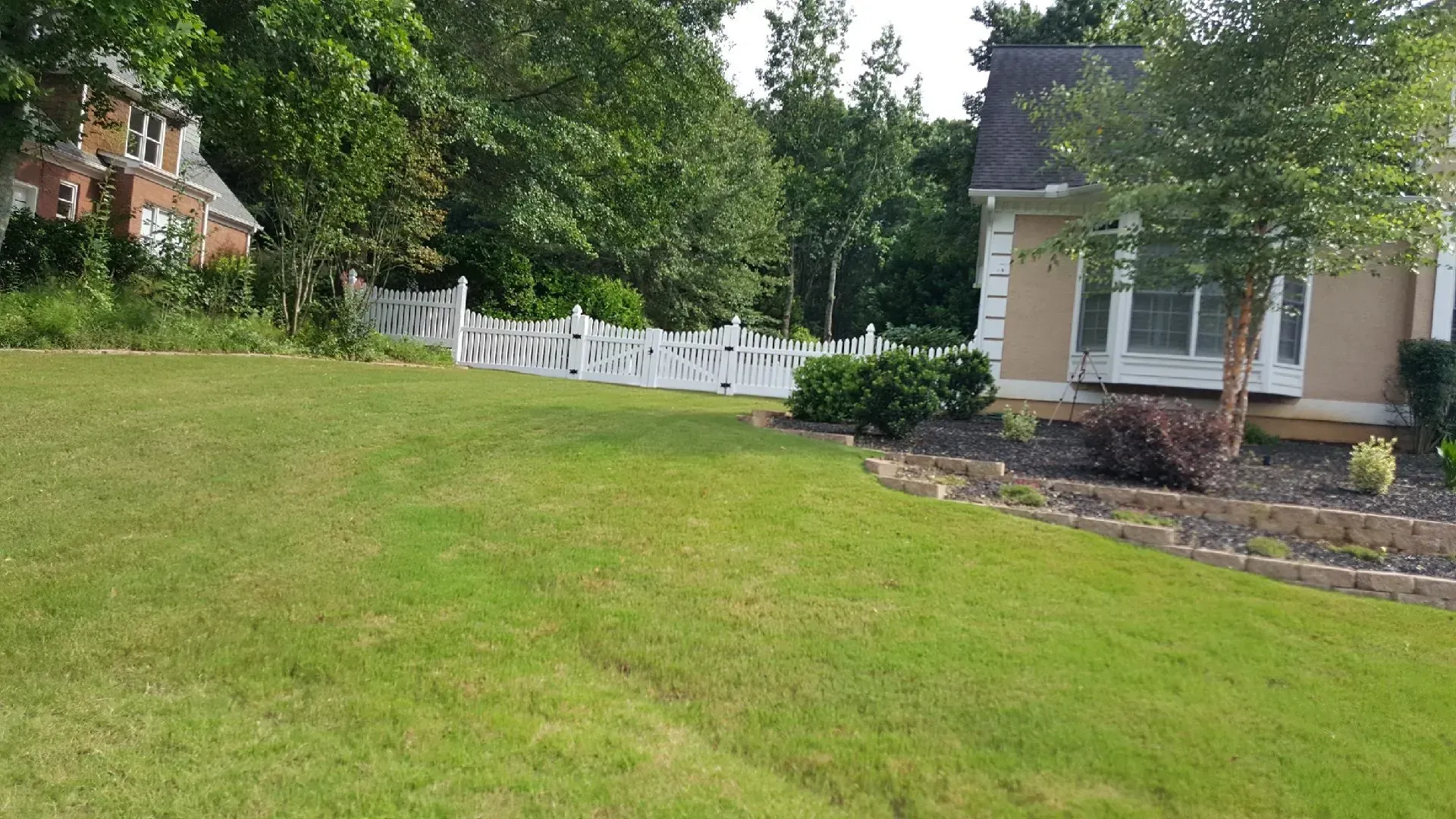 Lawn beside a house with white patio chairs, trees, and flower beds