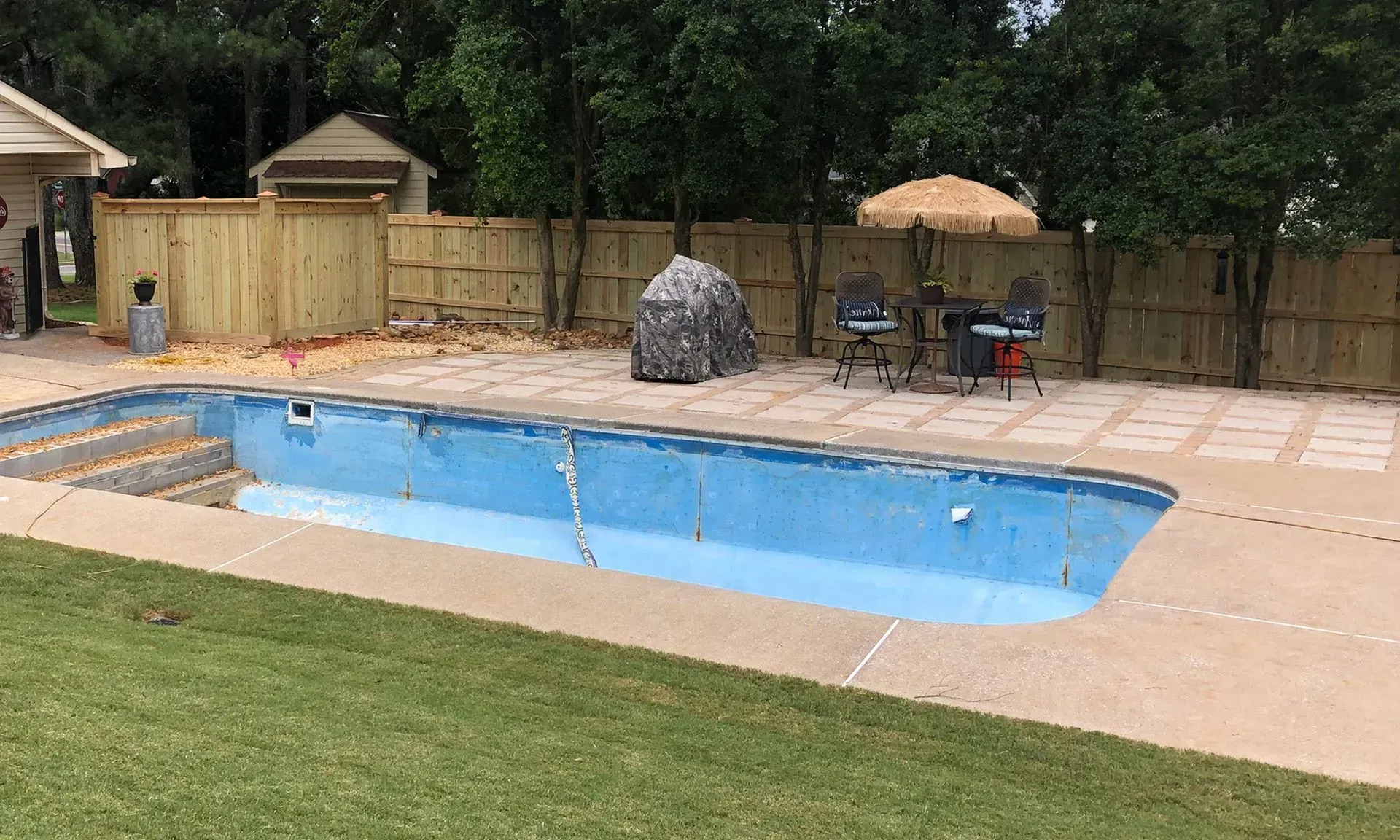 Backyard swimming pool with patio, umbrella table, fence, and trees surrounding it